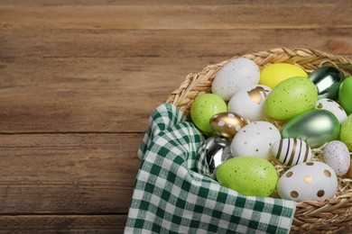 Many beautifully decorated Easter eggs in wicker basket on wooden table, space for text