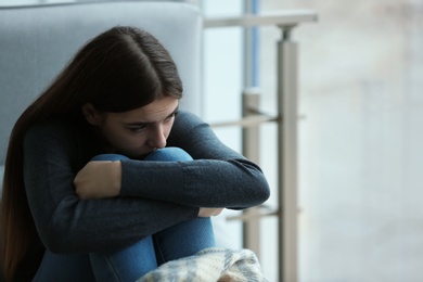 Photo of Upset teenage girl sitting at window indoors. Space for text