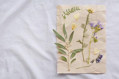 Sheet of paper with dried flowers and leaves on white fabric, top view. Space for text