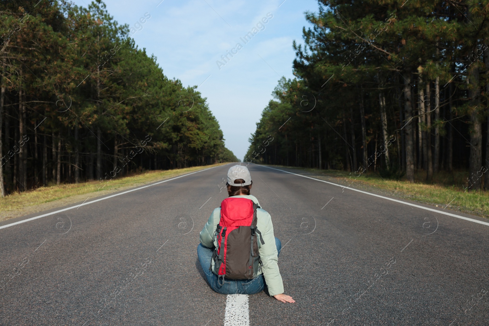 Photo of Young woman with backpack sitting on road near forest, back view