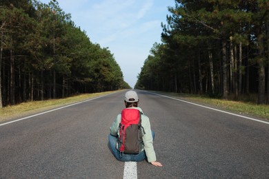 Young woman with backpack sitting on road near forest, back view