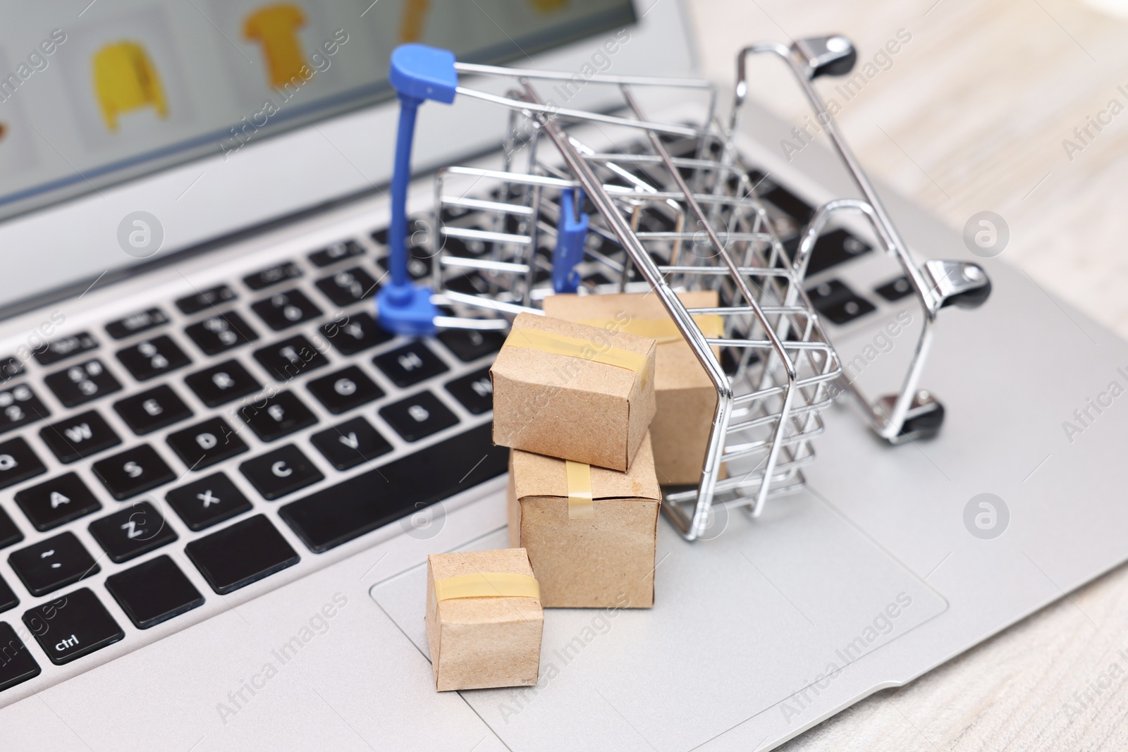 Photo of Internet store. Small cardboard boxes, shopping cart and laptop on white wooden table, closeup