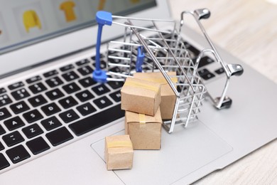 Internet store. Small cardboard boxes, shopping cart and laptop on white wooden table, closeup