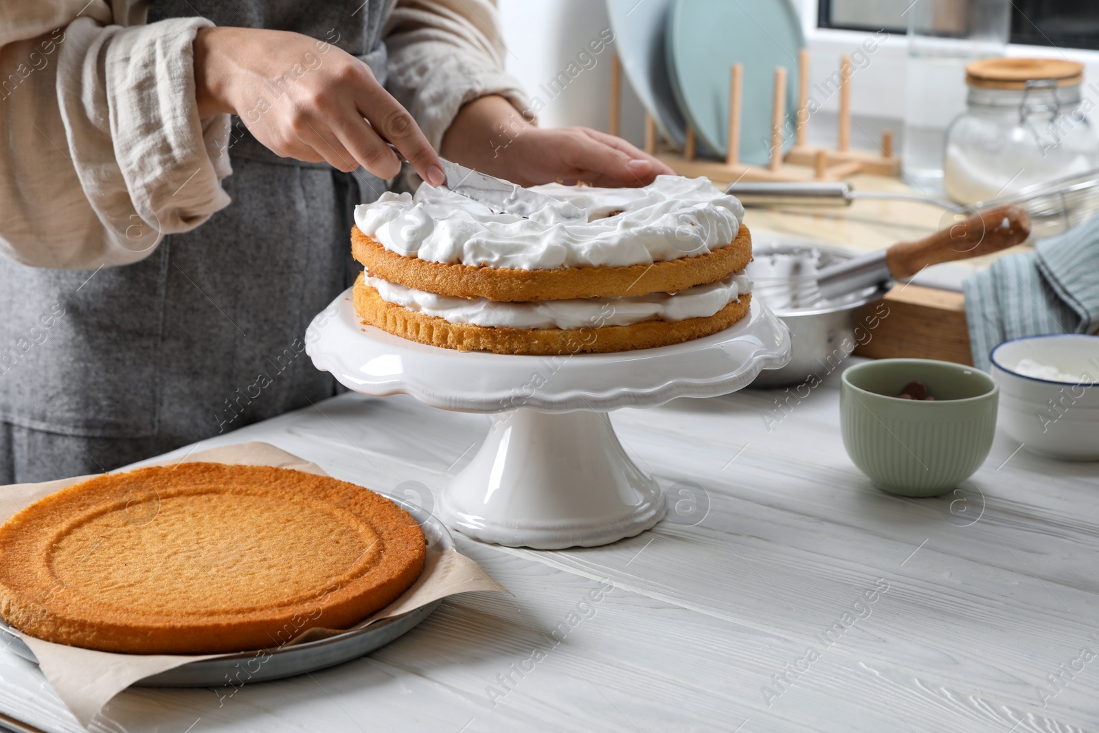 Photo of Woman smearing sponge cake with cream at white wooden table in kitchen, closeup