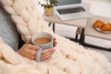 Woman wrapped in soft knitted blanket holding cup of coffee indoors, closeup