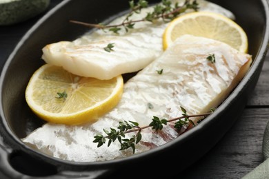 Fresh raw cod fillets with thyme and lemon in baking dish on black wooden table, closeup
