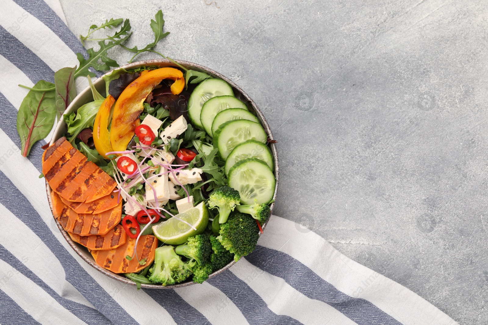 Photo of Bowl with many different vegetables and tofu on grey table, top view and space for text. Vegan diet