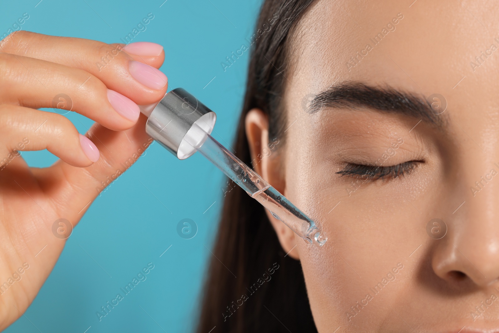 Photo of Young woman applying serum onto her face on light blue background, closeup