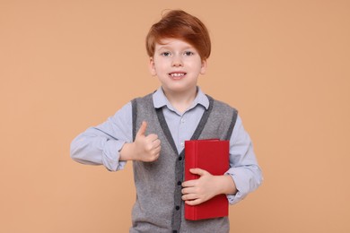 Photo of Smiling schoolboy with book showing thumb up on beige background