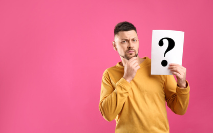 Emotional man holding paper with question mark on pink background, space for text