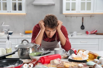 Photo of Upset man in messy kitchen. Many dirty dishware, utensils and food leftovers on table