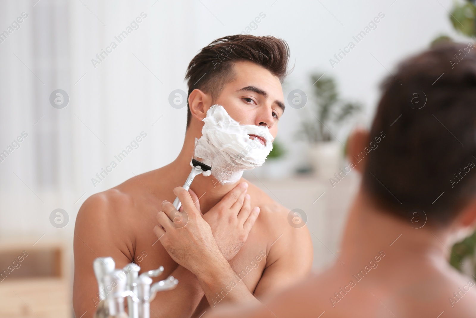 Photo of Young man shaving near mirror in bathroom