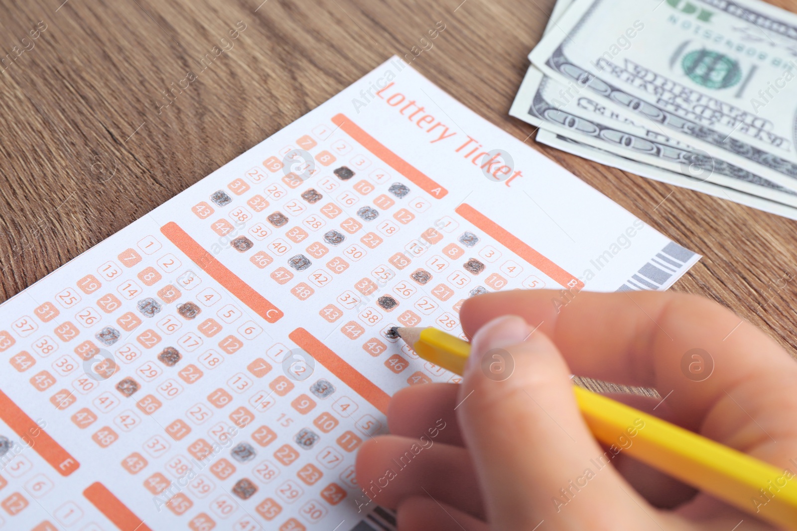 Photo of Woman filling out lottery ticket with pencil and money on wooden table, closeup