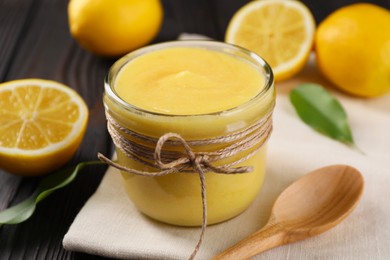 Delicious lemon curd in glass jar, fresh citrus fruits and spoon on wooden table, closeup