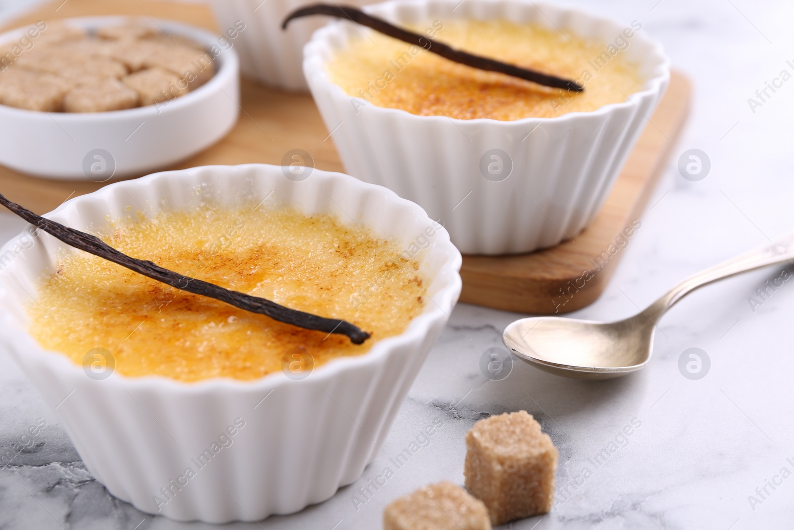 Photo of Delicious creme brulee in bowls, vanilla pods, sugar cubes and spoon on white marble table, closeup