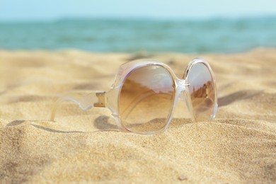 Photo of Stylish sunglasses on sandy beach near sea, closeup