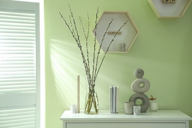 Photo of Glass vase with pussy willow tree branches and decor on white table near light green wall indoors