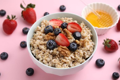 Photo of Tasty oatmeal with strawberries, blueberries and walnuts in bowl surrounded by fresh berries on pink background