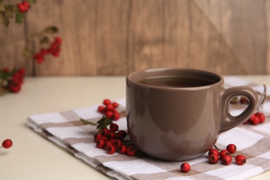 Photo of Brown cup with hawthorn tea and berries on beige table, closeup. Space for text