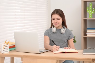 Photo of Cute girl with headphones writing in notepad near laptop at desk in room. Home workplace