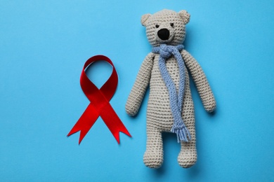 Cute knitted toy bear and red ribbon on blue background, flat lay. AIDS disease awareness