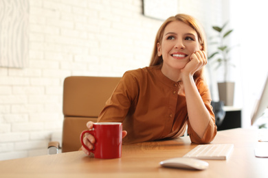 Young woman with cup of drink relaxing at table in office during break