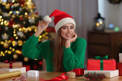Photo of Beautiful young woman in Santa Claus hat with Christmas gifts and decor at table in room
