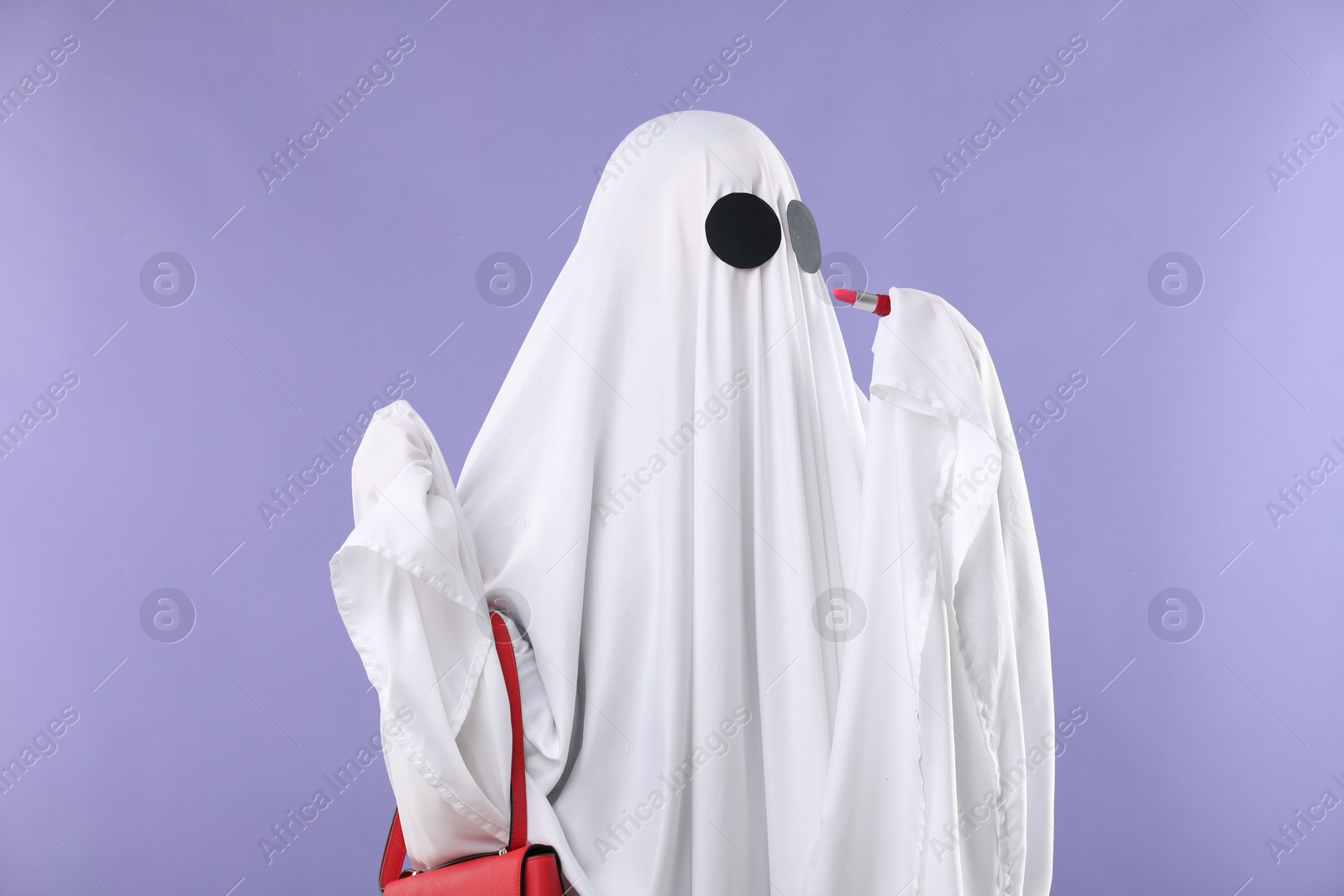 Photo of Glamorous ghost. Woman in white sheet with stylish bag and lipstick on violet background