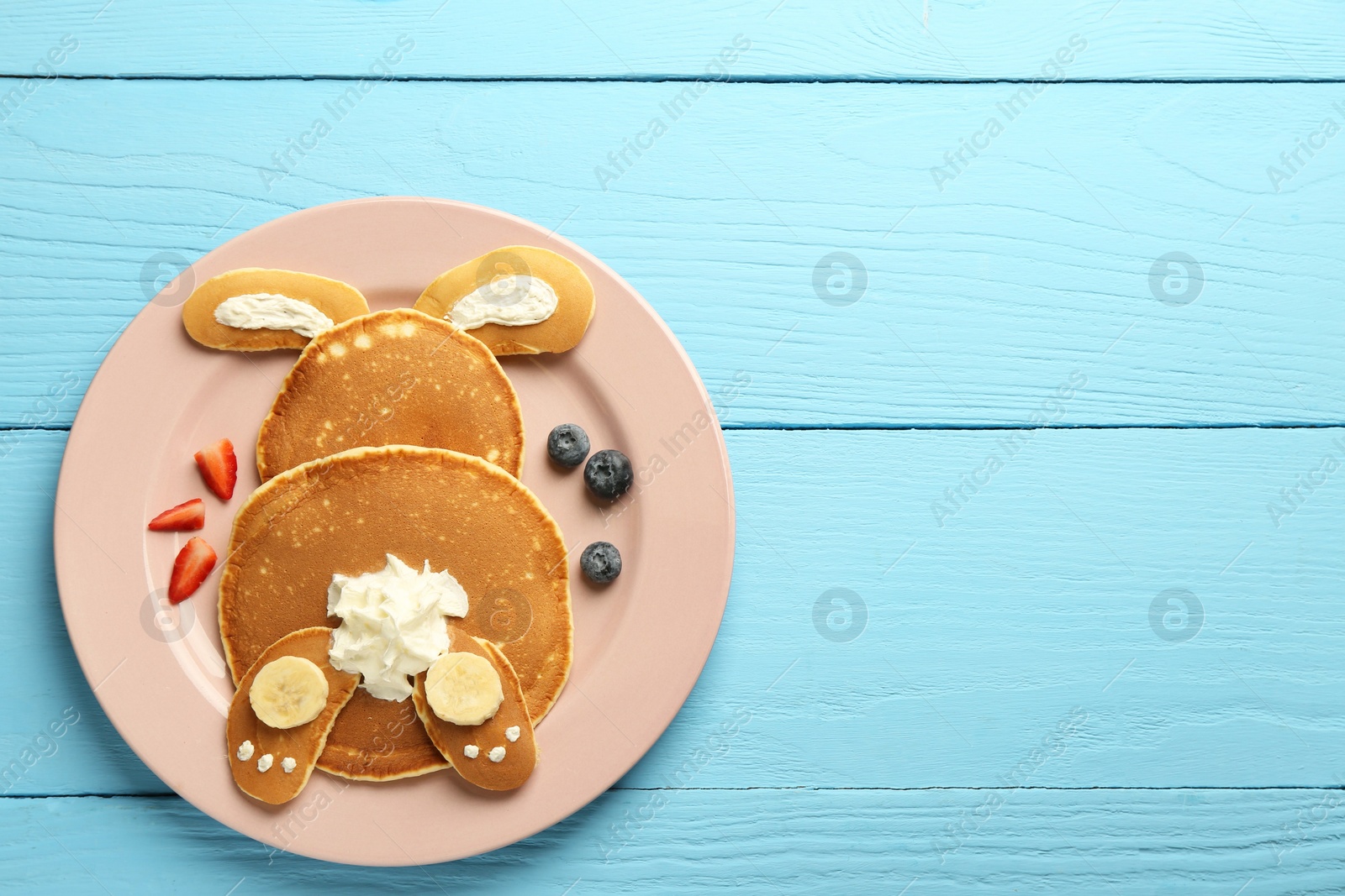 Photo of Creative serving for kids. Plate with cute bunny made of pancakes, berries, cream and banana on light blue wooden table, top view. Space for text