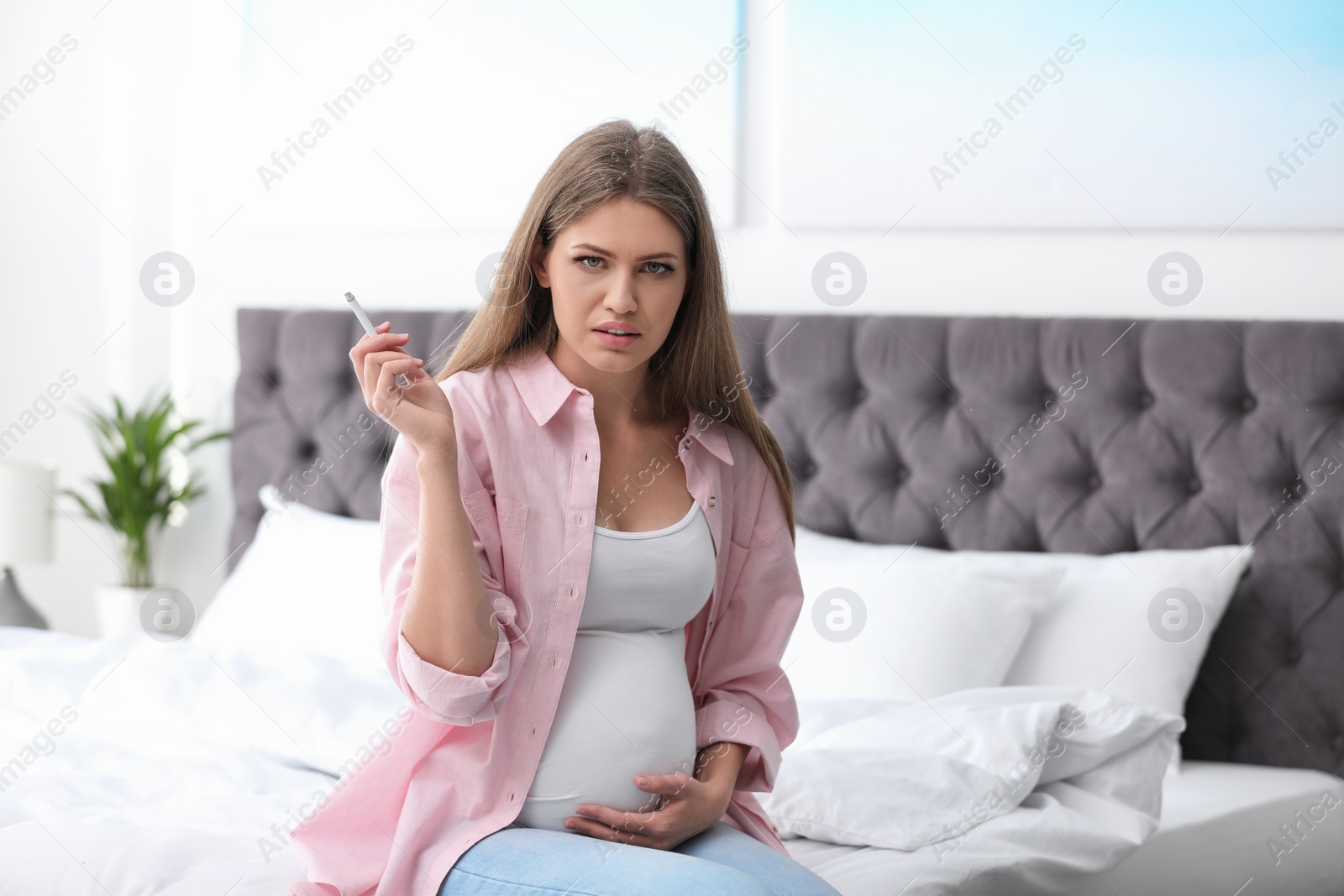 Photo of Young pregnant woman smoking cigarette in bedroom. Harm to unborn baby