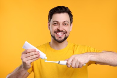 Photo of Happy man squeezing toothpaste from tube onto electric toothbrush on yellow background