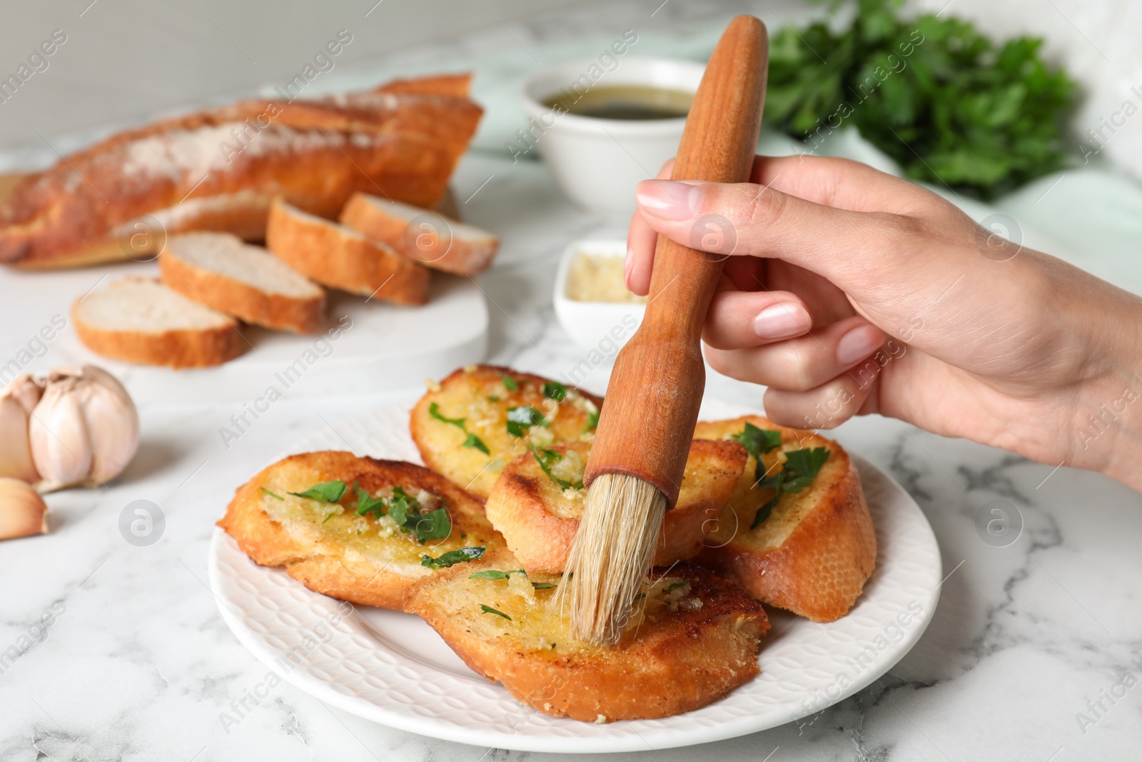 Photo of Woman brushing slices of delicious toasted bread with garlic and herbs on white marble table, closeup