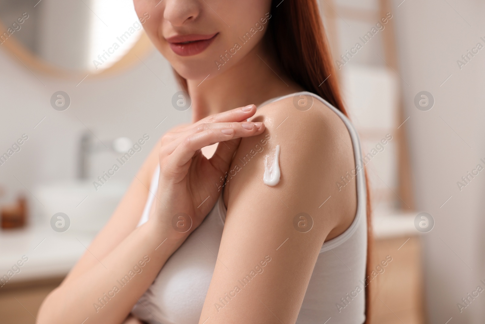 Photo of Young woman applying body cream onto shoulder in bathroom, closeup