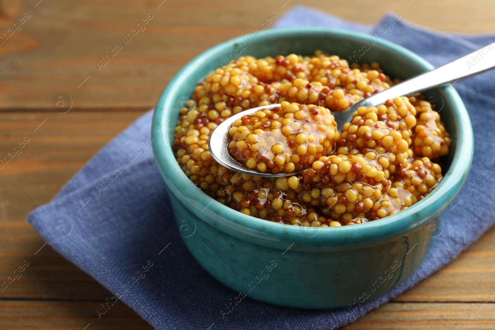 Photo of Whole grain mustard in bowl and spoon on wooden table, closeup