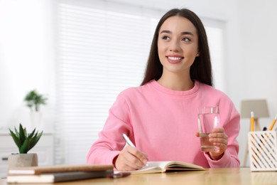 Photo of Young woman with glass of water writing in notebook at wooden table indoors