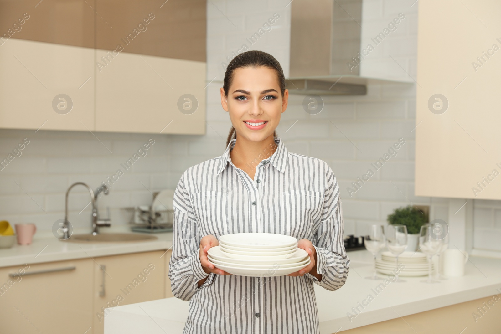 Photo of Beautiful young woman holding stack of clean dishes in kitchen