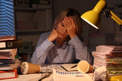 Overwhelmed woman surrounded by documents and paper coffee cups at table in office at night