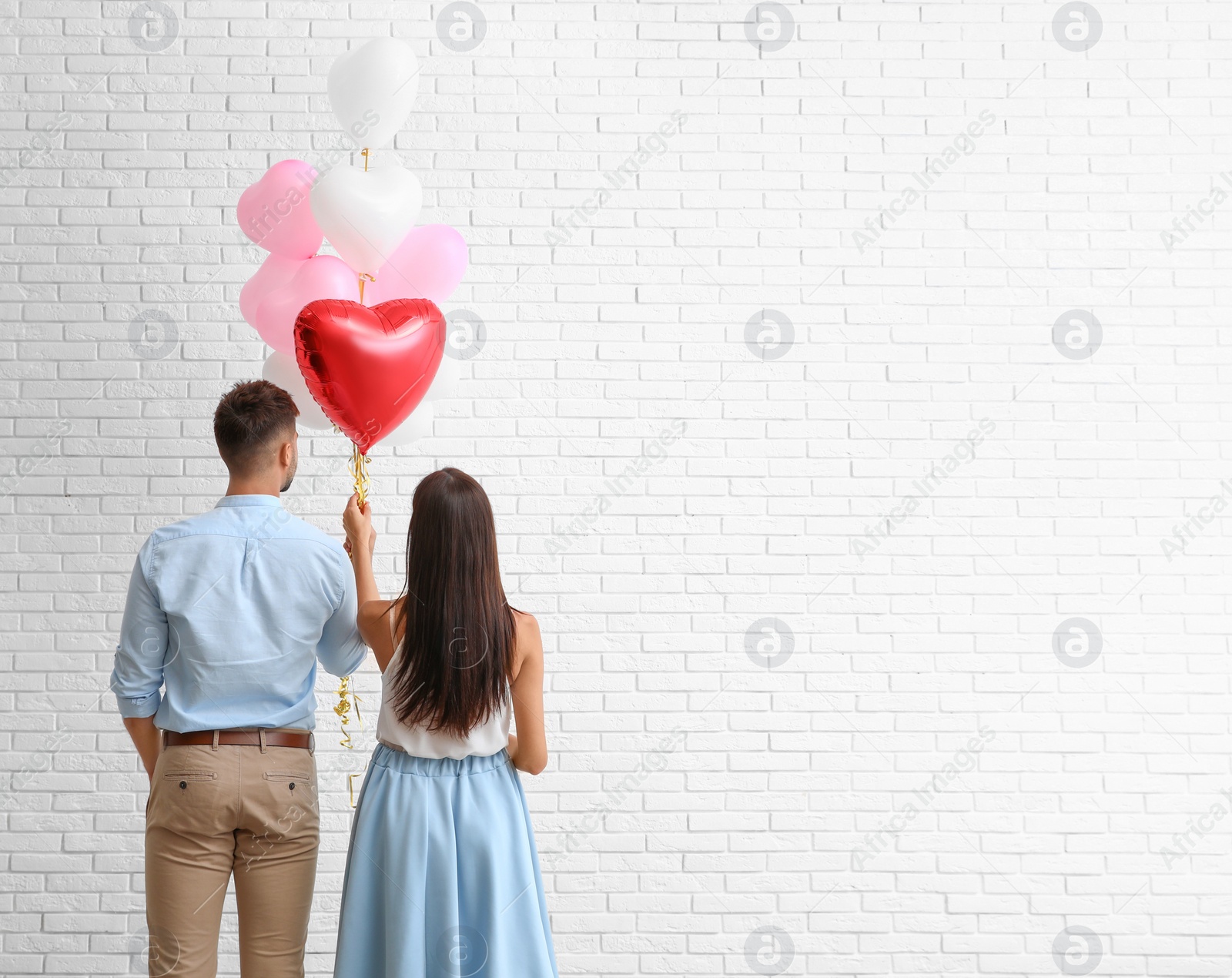 Photo of Young couple with air balloons near white brick wall. Celebration of Saint Valentine's Day