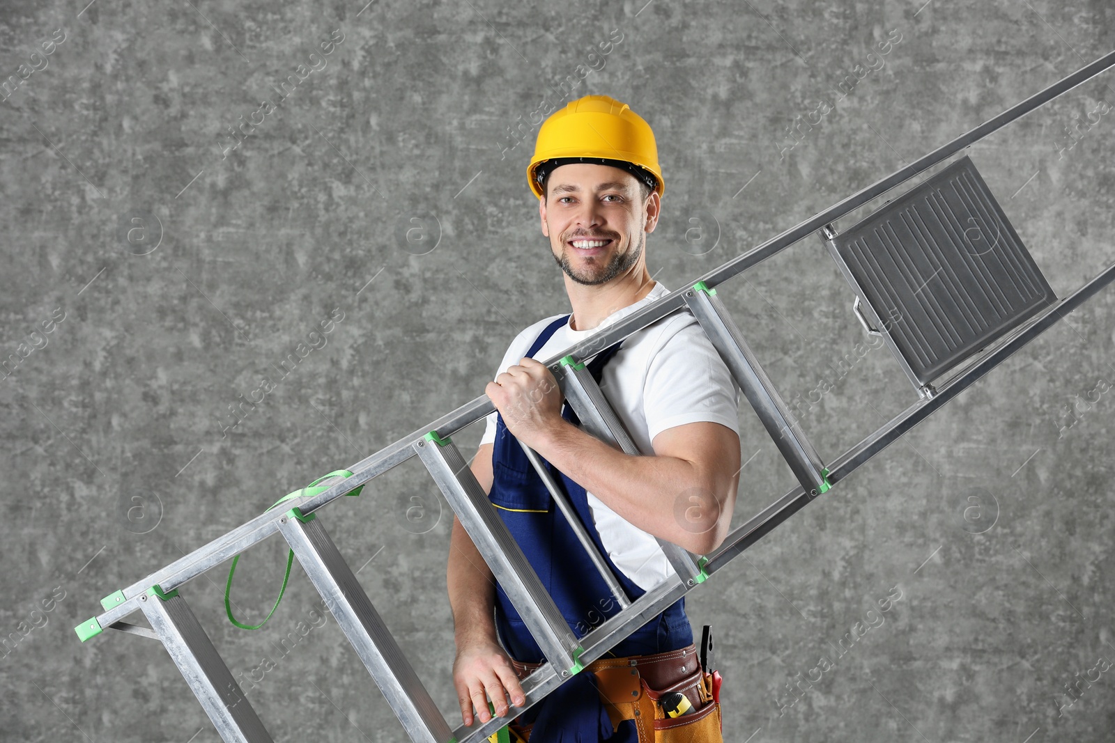 Photo of Handsome working man in hard hat holding ladder against color background