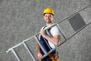 Handsome working man in hard hat holding ladder against color background