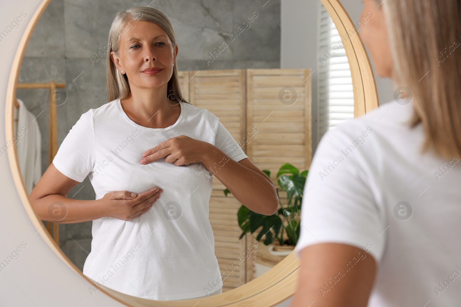 Photo of Beautiful senior woman doing breast self-examination near mirror in bathroom