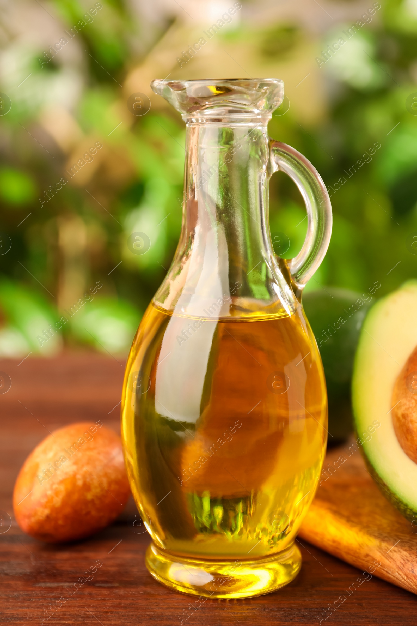 Photo of Fresh avocados and jug of cooking oil on wooden table against blurred green background, closeup