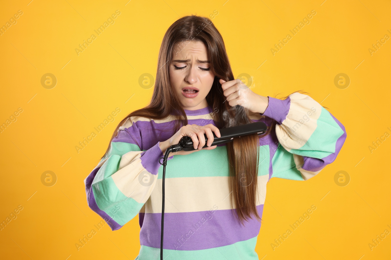 Photo of Stressed young woman with flattening iron on yellow background. Hair damage