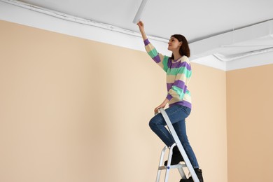 Photo of Young woman installing ceiling lamp on stepladder indoors, space for text