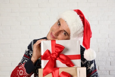 Photo of Happy man in Christmas sweater and Santa hat holding gift boxes near white brick wall