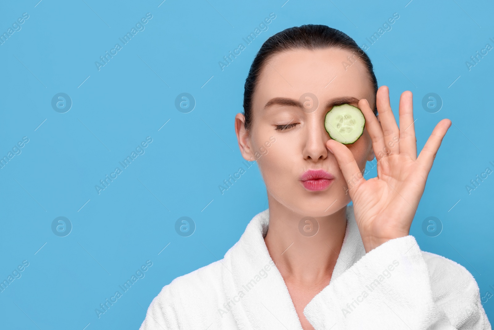 Photo of Beautiful woman covering eye with piece of cucumber and sending air kiss on light blue background, space for text