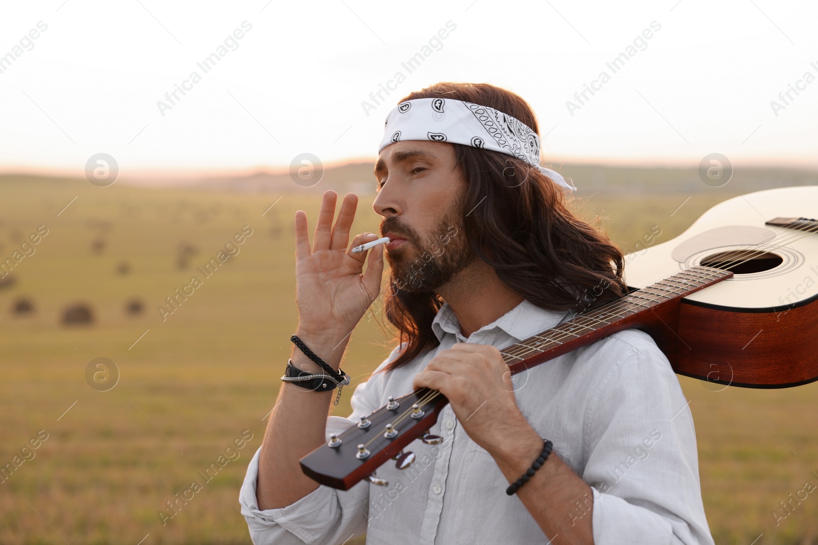 Photo of Stylish hippie man with guitar smoking joint in field, space for text