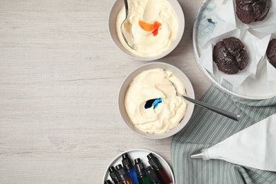 Bowls of different cream with food coloring, bottles of bright liquid and cupcakes on white wooden table, flat lay. Space for text