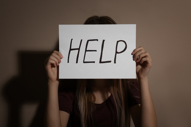 Photo of Young woman with sign HELP near beige wall. Domestic violence concept