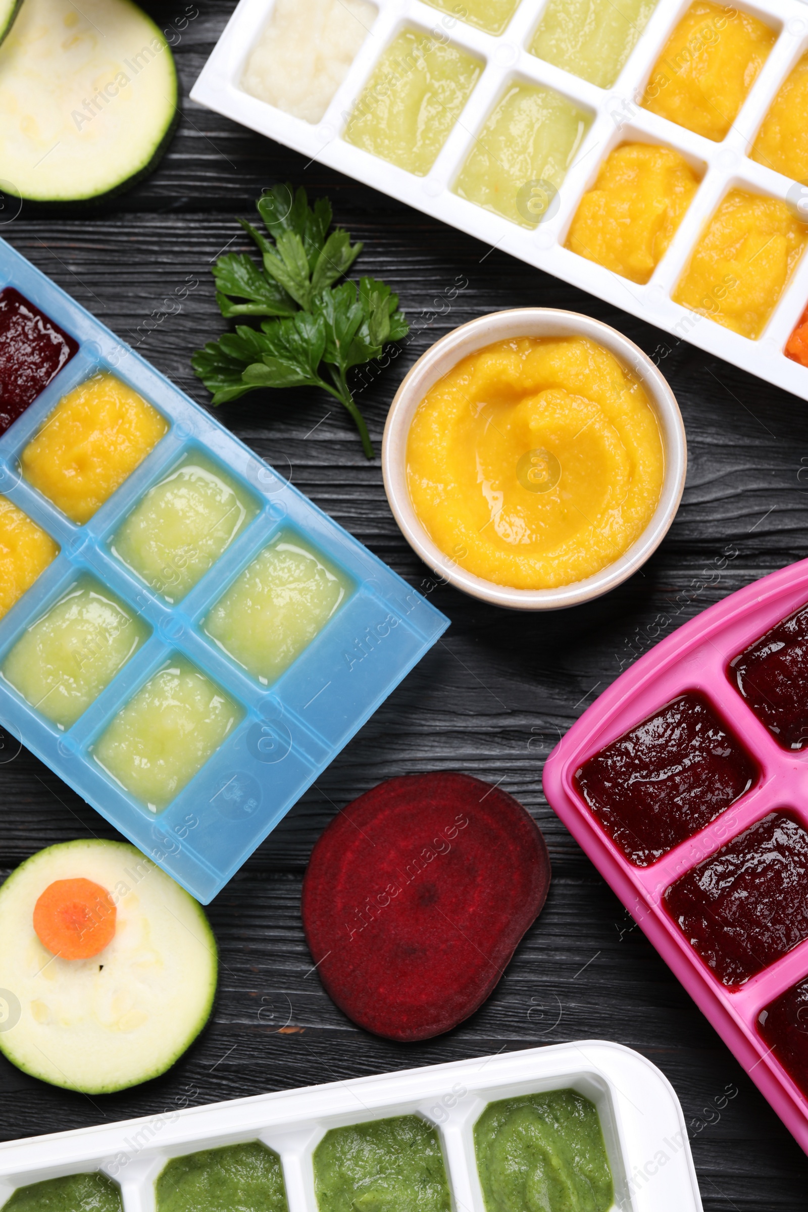 Photo of Different frozen purees in ice cube trays and ingredients of black wooden table, flat lay. Ready for freezing